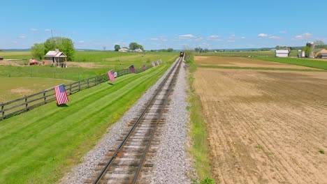 An-Aerial-View-of-a-Steam-Train-Approaching-Slowly-Thru-Farmlands-and-a-Fence-with-Many-American-Flags-Waving-in-the-Wind-on-a-Sunny-Spring-Day