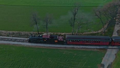 An-Aerial-Side-View-of-a-Steam-Passenger-Train-Traveling-Blowing-Smoke,-on-a-Single-Track,-Thru-Farmlands-on-a-Sunny-Spring-Day