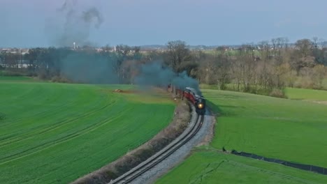 An-Aerial-View-of-a-Steam-Passenger-Train-Traveling-Blowing-Smoke,-on-a-Single-Track,-Coming-Around-a-Curve-on-a-Sunny-Spring-Day