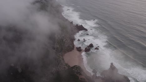 Aerial-view-of-Praia-da-ursa-Portugal-with-low-clouds-above-the-sea,-drone-shot