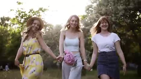 Women-friendship.-Three-attractive-girls-in-summer-outfits.-Holding-hands-together,-walking-playfully-in-the-forest.-Happy,-smiling