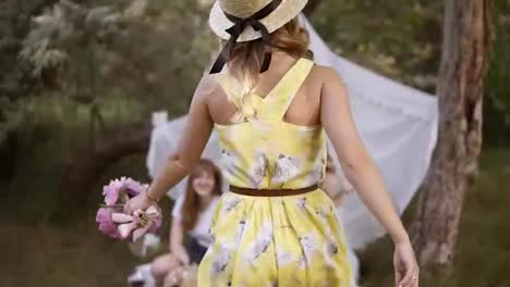 Hen-party-in-the-green-garden.-Girls-friends-are-meeting-their-friend.-Girl-in-yellow-dress-and-summer-hat-happily-coming-to-a-picnic-with-friends