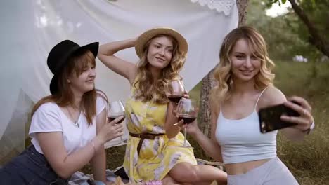 Three-beautiful,-modern-women-sitting-on-a-grass-with-glasses-of-red-wine.-One-blonde-girl-taking-a-selfie-with-her-girlfriends.-Picnic-outdoors.-Hen-party