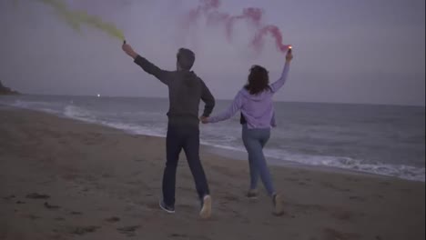 Back-view-of-young-couple-running-with-colored-smoke-grenade-in-their-hands-by-the-sea-during-sunset.-Fun,-colorful-smoke-around-them