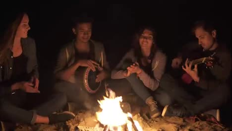 Multiracial-group-of-young-women-and-men-sitting-by-the-bonfire-late-at-night-and-singing-songs,-playing-guitar-and-percussion