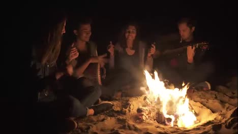 Two-young-women-and-two-men-sitting-by-the-bonfire-late-at-night-and-singing-songs,-playing-guitar-and-percussion