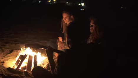 Round-camera-movement:-multiracial-group-of-young-people-sitting-by-the-bonfire-late-at-night-and-singing-songs,-playing-guitar-and-percussion