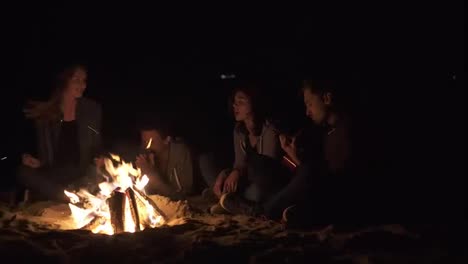Multiracial-group-of-young-boys-and-girls-sitting-by-the-bonfire-late-at-night-and-singing-songs-and-playing-guitar