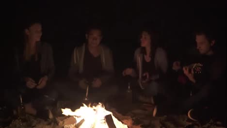 Closeup-view-of-the-bonfire-late-at-night.-Young-people-sitting-by-the-fire-in-the-evening,-playing-guitar.-Cheerful-friends-singing-songs,-talking-and-having-fun-together