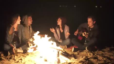 Camera-moves-around-the-bonfire-late-at-night:-young-cheerful-people-sitting-by-the-fire-in-the-evening,-playing-guitar-and-drinking-beer.-Cheerful-friends-singing-songs,-talking-and-having-fun