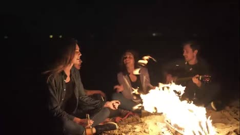 Young-cheerful-people-singing-songs-by-the-bonfire-late-at-night,-playing-guitar,-drinking-beer.-Cheerful-friends-talking-and-having-fun
