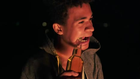 Young-attractive-african-american-opening-a-beer-bottle-with-teeth-and-starting-to-drink-it,-looking-in-the-camera-while-sitting-by-the-bonfire-late-at-night