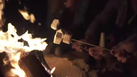 Closeup-view-of-hands-holding-sticks-with-marshmallows-and-frying-them-at-night.-Group-of-people-sitting-by-the-fire-late-at-night