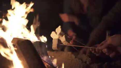 Closeup-view-of-marshmallow-on-sticks-being-fryed-by-the-bonfire.-Group-of-people-sitting-by-the-fire-late-at-night