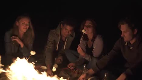 Young-cheerful-friends-sitting-by-the-fire-on-the-beach-late-at-night,-roasting-marshmallow-on-sticks-over-the-fire-together