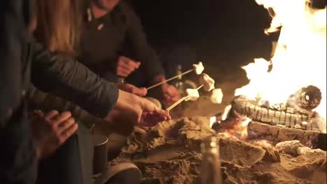 Group-of-young-multiethnic-people-sitting-by-the-fire-on-the-beach-late-at-night,-roasting-marshmallow-on-sticks-over-the-fire-together