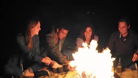Group-of-young-multiethnic-people-roasting-marshmallow-on-sticks-over-the-fire-together-on-the-beach-late-at-night,-drinking-beer