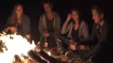 Group-of-young-friends-sitting-by-the-fire-late-at-night,-grilling-sausages-and-drinking-beer,-talking-and-having-fun