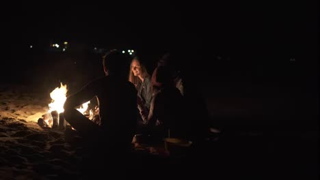 Back-view-of-young-diverse-people-sitting-together-by-the-fire-late-at-night.-Cheerful-friends-talking-and-having-fun-together,-drinking-beer