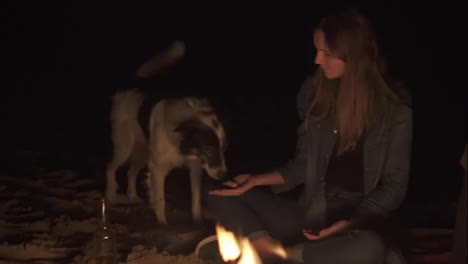 Attractive-caucasian-woman-feeding-cute-dog-sitting-by-the-bonfire-late-at-night.-Closeup-of-kind-woman's-hand-with-piece-of-sausage-and-face-of-playful-dog