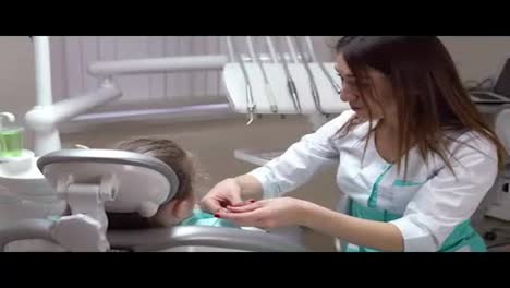 Friendly-female-dentist-putting-a-napkin-on-the-cute-little-girl,-taking-on-gloves-and-protective-glasses-before-doing-routine-check-up.-Shot-in-4k