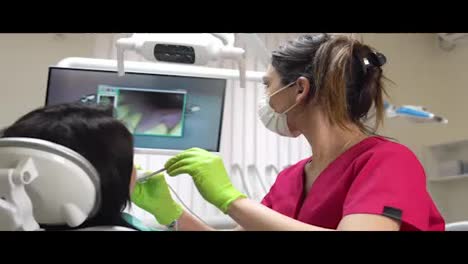 Young-female-dentist-examining-the-mouth-of-a-patient-with-an-intraoral-camera-and-showing-image-on-the-screen.-Shot-in-4k