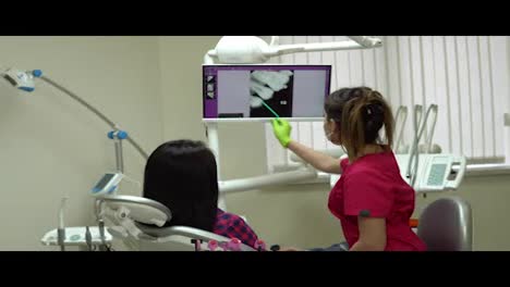 Young-female-doctor-pointing-on-the-screen-with-x-ray,-explaining-her-patient-needed-treatment.-Young-woman-sitting-in-the-dentist-chair-and-looking-at-the-monitor.-Shot-in-4k