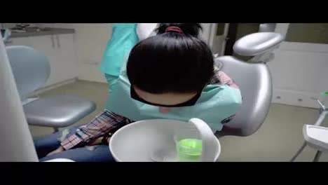 Junge-Frau-Mit-Roter-Schutzbrille-Spült-Sich-Während-Der-Behandlung-Beim-Zahnarzt-Den-Mund-Aus.-Glas-Mit-Grüner-Flüssigkeit-In-Der-Hand.-In-4k-Gedreht