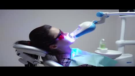 Young-woman-with-an-expander-in-mouth-and-red-protective-glasses-getting-UV-whitening-at-the-dentist's-office-by-an-ultra-violet-machine.