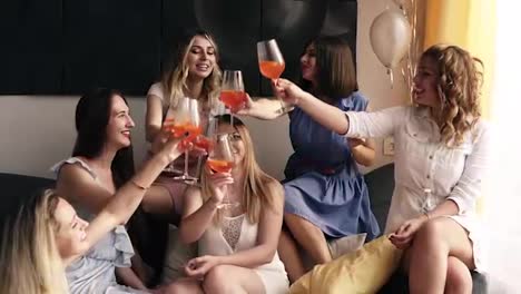 Group-of-gorgeous-young-women-celebrating-together.-Sitting-on-a-couch-in-modern,-fancy-interior.-Clinking-and-raising-hands-with-cocktails.-Posing-with-glasses.-Elegant-casual-clothes.-Slow-motion