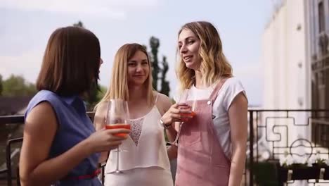 Three-modern,-young-women-are-talking-and-drinking-orange-cocktails-from-wine-glasses.-Sharing-stories-and-laughing.-Hanging-out-on-a-terrace-on-daytime.-Slow-motion
