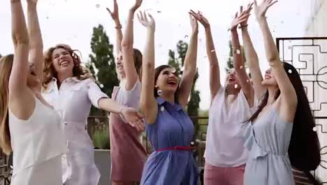 Group-of-young-girls-dressed-in-casual-partying-outdoors-on-terrace-silver-confetti-in-the-air-at-daytime-during-their-carefree-hen-party.-Dancing-and-hugging-each-other-in-circle.-Slow-motion