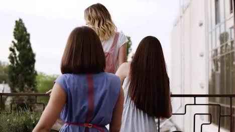 Women's-friendship.-The-blonde-girl-is-standing-near-balcony-railings-and-looking-forward.-Girlfriends-are-coming-to-her-one-by-one-and-hugging.-Blurred-trees-in-perspective.-Backside-view