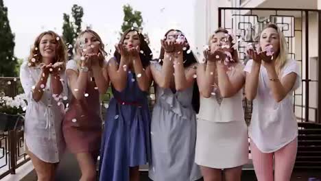 Front-view-of-six-attractive-caucasian-young-women-playfully-blowing-bright-colored-confetti-from-their-hands-together.-stylish-clothes.-Bachelorette-party.-slow-motion