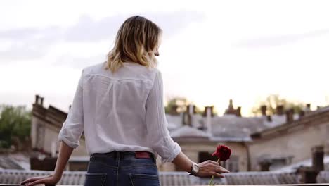 Sensual,-slender-woman-in-jeans-and-white-shirt-is-standing-on-a-balcony-with-red-flower-in-her-hand.-Blonde-girl.-Beautiful-town-perspective.-Backside