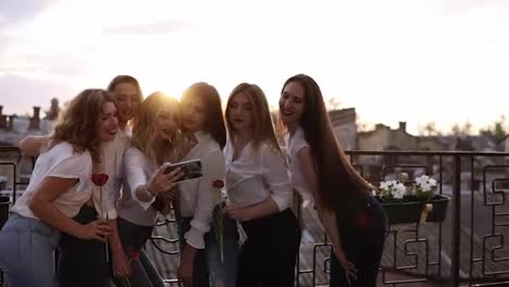 Emotional-bridesmaid-and-attractive-girlfriends-talking-selfie-with-mobile-phone-and-smiling.-Caucasian-girls-in-white-shitrs-posing-and-holding-flowers-in-hands.-Outdoors.-Front-view