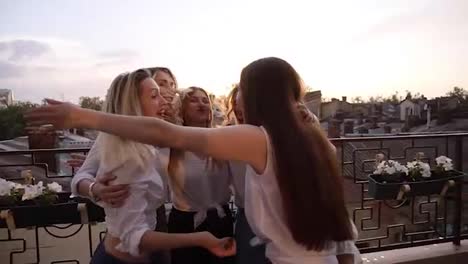 Happy,-young-girls-embraced-in-circle,-side-view.-Six-attractive-girls-hugging,-looking-each-other,-standing-outdoors-on-a-terrace.-Laughing.-Town-view-on-the-background