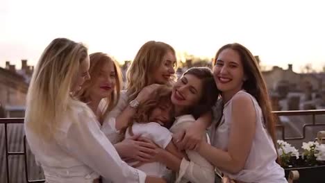 Attractive-caucasian-girls-are-hugging-standing-outside-on-a-terrace-or-balcony.-Six-beautiful-young-women-in-white-shirts-and-with-red-lipstick.-Hen-party.-Evening-dusk.-Love-and-friendship