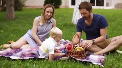 Happy-family-sitting-on-a-plaid-at-the-picnic-on-the-green-beautiful-meadow-on-a-sunny-day.-Their-son-is-sitting-near.-Enjoy-the-picnic.-Outdoors