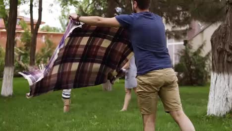Father-in-brown-shorts-and-blue-shirt-is-spreading-the-plaid-for-picnic.-Happy-mother-is-playing-with-her-little-son-on-the-blurred-background.-Green-grass.-Slow-motion