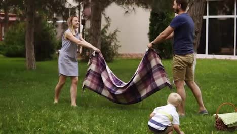 Slow-motion-of-a-young-family-with-a-son-on-a-green-lawn.-Parents-are-spreading-the-plaid.-Excitement.-Blonde-little-boy-on-a-forward-perspective
