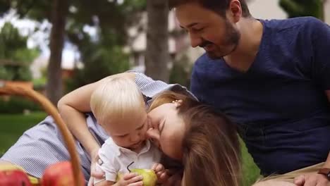 Close-up-of-a-young-parents-sitting-close-to-each-other-with-their-little-son.-Picnic-outdoors-on-a-plaid.-Picnic-basket-with-apples.-Smiling,-happy-family
