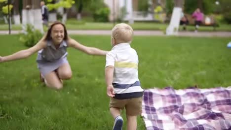 Little-blond-boy-running-to-his-mother.-Mom-opened-her-hands-and-smiling-catches-the-baby.-Happy-mother,-loving-son.-Picnic-outdoors.-Green-park.-Slow-motion