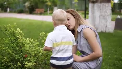 A-little-boy-is-interested-in-a-green-bush.-Touching,-sniffing-it.-Mom-talks-to-her-son,-explains.-Green-park.-Slow-motion