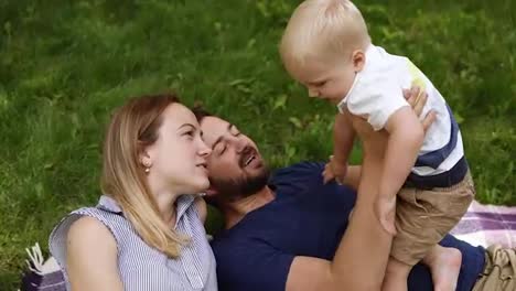 High-angle-footage-of-beautiful-family-of-three.-Lying-on-green-grass,-playing-with-his-little-son.-Happy-lifestyle.-Sunny-day