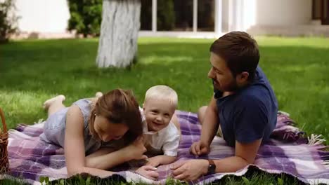 Family-picnic-concept.-Young-parents-lying-on-a-grass,-playing-with-his-son.-Have-fun.-Child-with-fair-hair-climbs-to-his-father's-back.-Embraces-his-neck.-Laughing