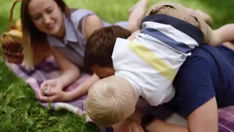 Caucasian-couple-have-a-good-time-oudoors-with-their-son.-Lying-on-a-plaid-in-the-park.-Picnic.-Father-plays-with-the-son.-Embrace,-kissing-him,-play-a-game.-Close-up