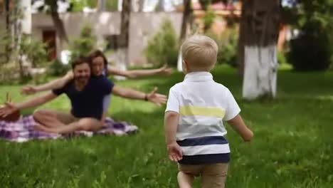 Family-idyll.-Happy-child-running-to-his-parents-in-a-blurred-perspective.-Close-up-of-young-couple-hugging-his-little-blonde-son-while-sitting-on-the-grass,-plaid.-Picnic-in-the-park.-Slow-motion