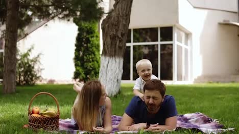 Happy-joyful-young-family-father,-mother-and-little-son-having-fun-outdoors,-playing-together-in-summer-park,-countryside.-Mom,-Dad-and-son-on-fathers-back-junping-and-laughing.-White-house-on-the-background.-Slow-motion