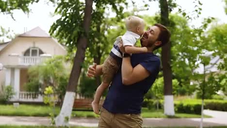 Young-father-with-beard-in-casual-clothes-is-holding-in-arms-his-little-blonde-son.-Boy-bare-feet.-Man-kissing-his-son.-Cottage-street-on-the-background.-Green-and-bright